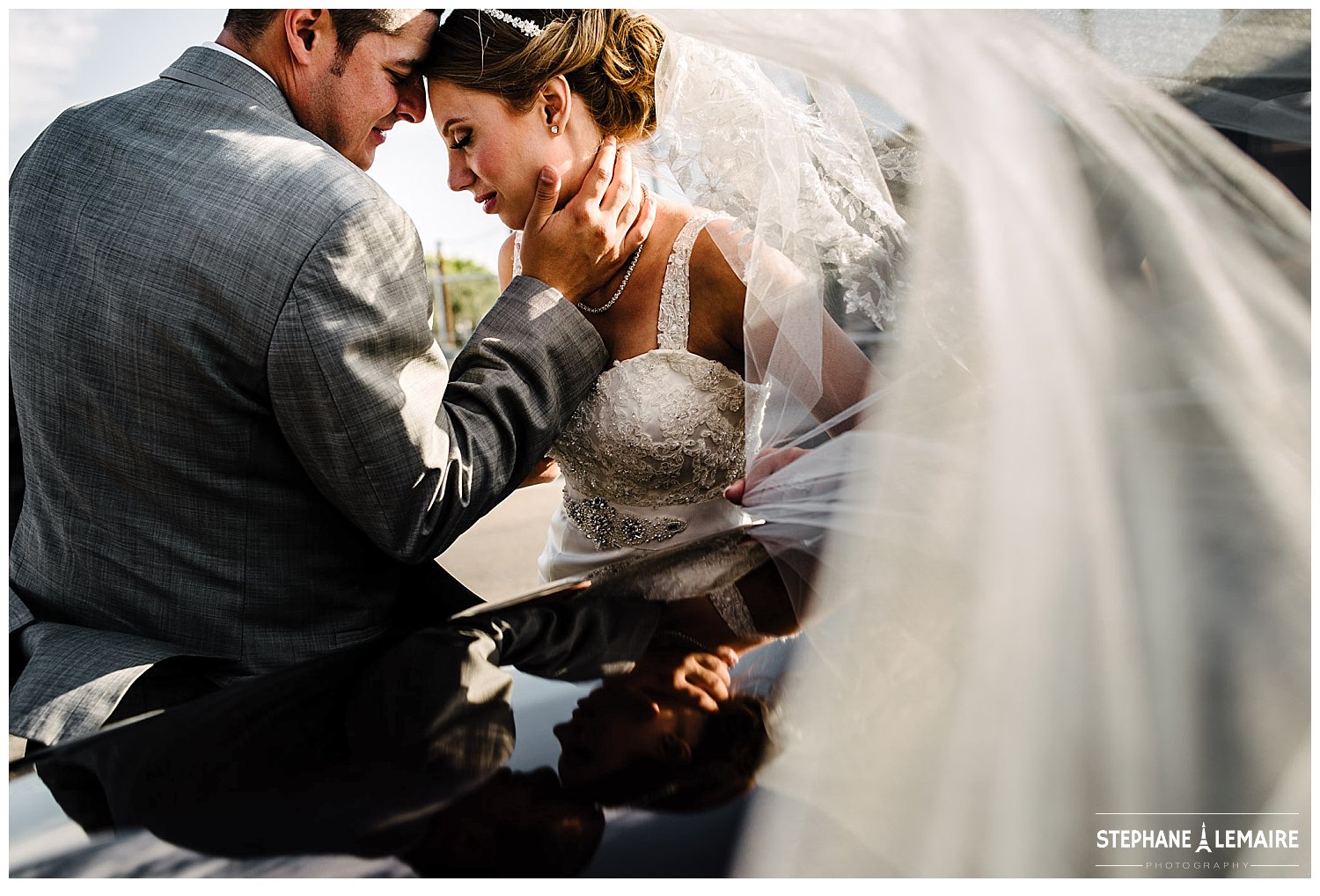 Bride and groom hugging next to vintage car with veil blowing in the wind.